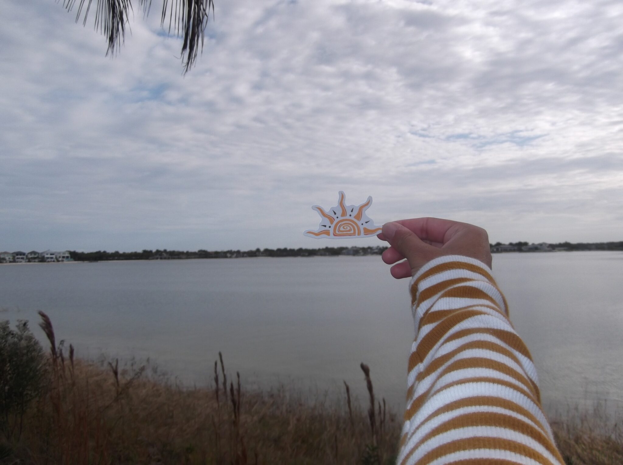 A hand holding up the Selah Studios sun sticker against a cloudy sky over a lake