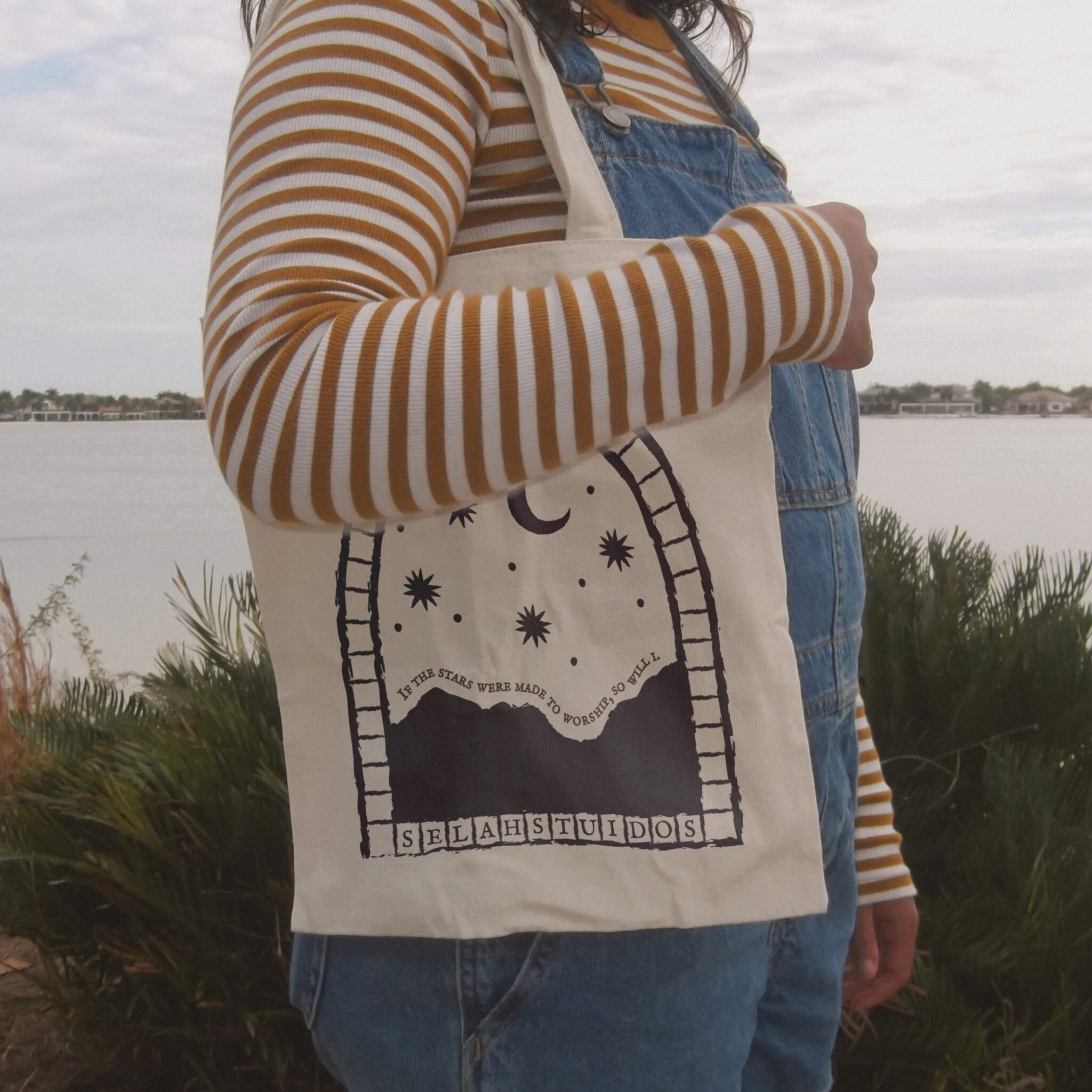 A person in a striped top carrying a cream tote bag printed with the arch-and-stars decal by the water